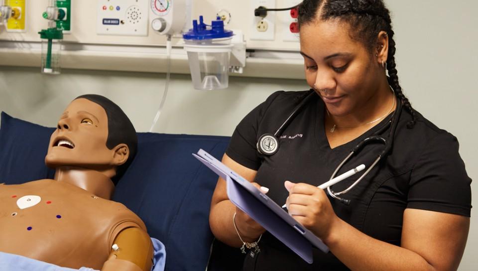 A physician assistant student wears a stethoscope and takes notes on a tablet while in a sim lab class