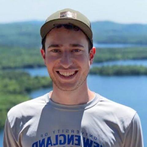 Mitchell Becker stands smiling at the camera atop a mountain. We see blue lakes and green mountains in the background